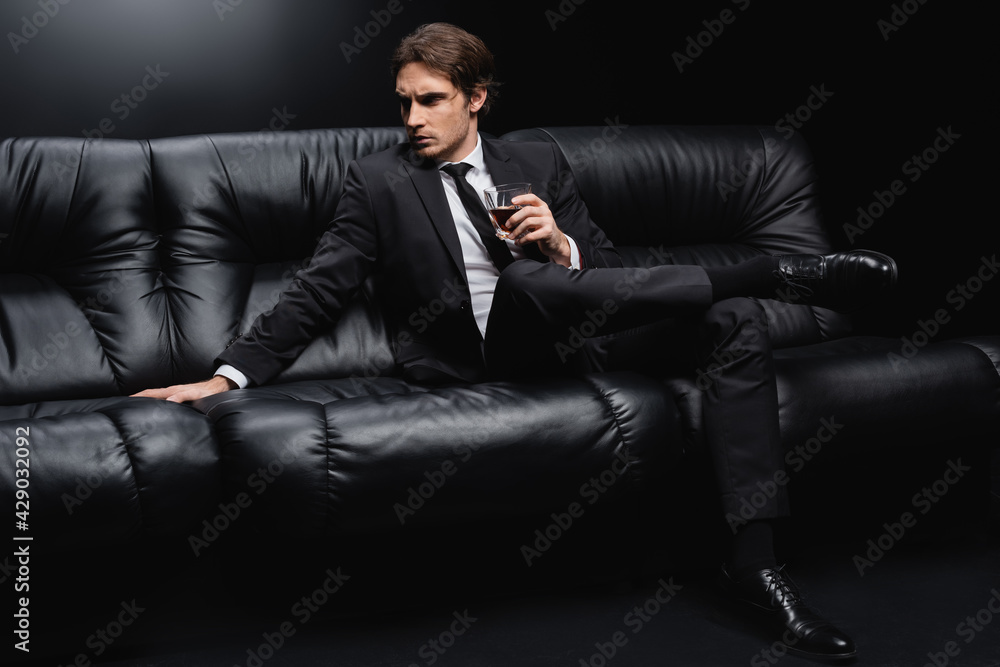 elegant man in suit sitting on leather couch with glass of whiskey on black background. elegant man in suit sitting on leather couch with glass of whiskey on black background.