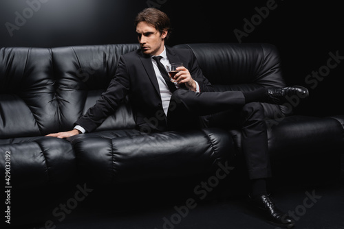 elegant man in suit sitting on leather couch with glass of whiskey on black background.