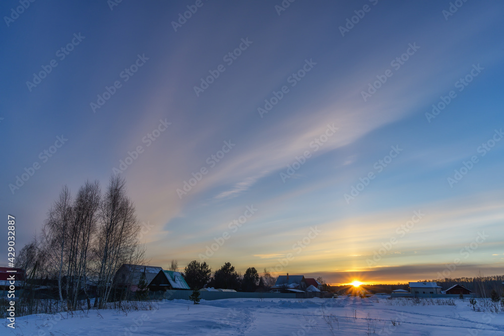 Sunset over the village on a frosty winter evening. The setting blue sky is decorated with large cirrus clouds painted in delicate pink tones. The ground is covered with pure white snow. Evening peace