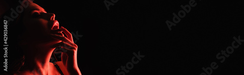 red lighting on seductive woman touching lips isolated on black, banner.