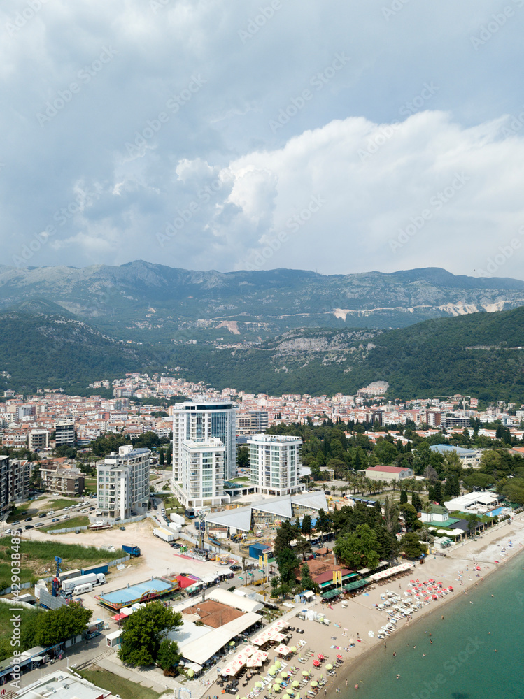 Budva, Montenegro - May 28 2018: Aerial view of city, surrounded by mountains, sandy beach, sea coastline, modern high-rise buildings. Umbrellas sun loungers on beach. Tourists sunbathing and swimming