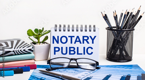 Against the background of a blue scheme and a white wall, black pencils in a stand, a flower, diaries and a notebook with the inscription NOTARY PUBLIC