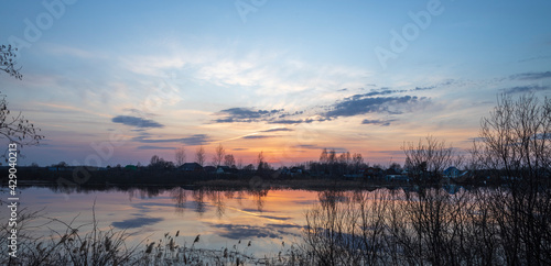 Evening landscape with a river. Village and forest on the horizon. Sunset on the river. The sky and evening clouds are reflected in the water. Branches of trees and bushes in the foreground. © Sergei