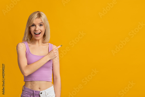 Look at this. Joyful blonde woman pointing finger aside at empty space, smiling at camera over yellow background