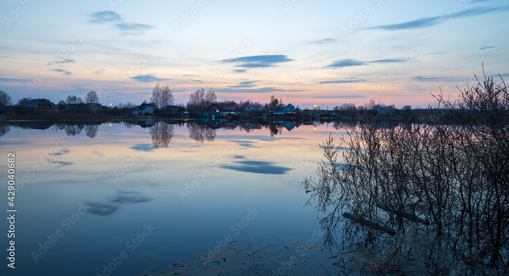 Evening landscape with a river. Village and forest on the horizon. Sunset on the river. The sky and evening clouds are reflected in the water. Branches of trees and bushes in the foreground.
