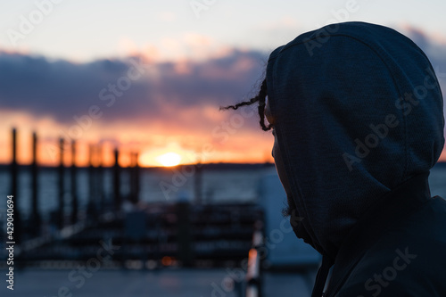 Silhouette of a man with braids and a hood watching the sunset by a dock on a lake