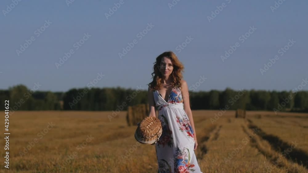 young attractive redhead woman in a white dress with a wicker basket walks in the field enjoying nature young attractive redhead woman in a white dress with a wicker basket walks in the field enjoying nature