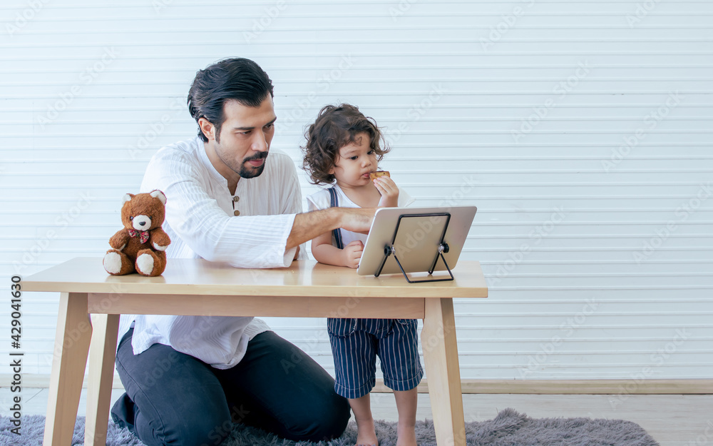 Caucasian handsome beard father or daddy taking care of his little curly hair daughter to study or learn online using tablet on wooden table when staying at home. Caucasian handsome beard father or daddy taking care of his little curly hair daughter to study or learn online using tablet on wooden table when staying at home.