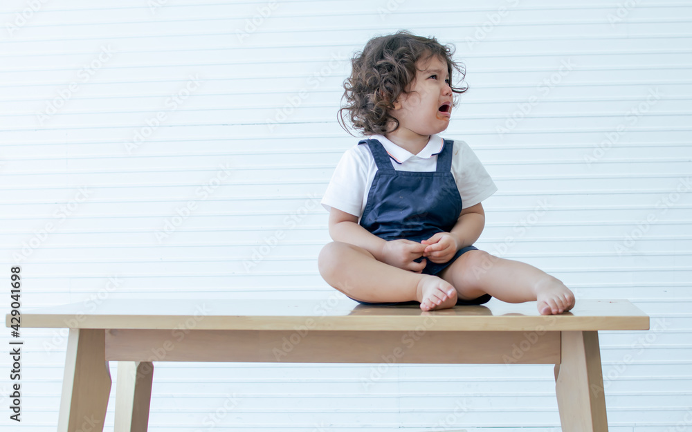 Caucasian little cute curly hair girl wearing white shirt, crying with sadness and sitting on wooden table in white living room at home. Caucasian little cute curly hair girl wearing white shirt, crying with sadness and sitting on wooden table in white living room at home.