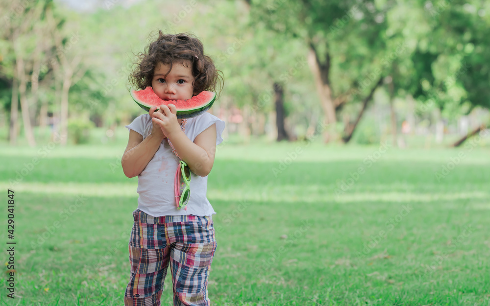Caucasian little cute girl standing, playing in outdoor green park for picnic, carrying, eating fruit, watermelon with freshness. Education, Friendship, Nature, Food Concept. Caucasian little cute girl standing, playing in outdoor green park for picnic, carrying, eating fruit, watermelon with freshness. Education, Friendship, Nature, Food Concept.