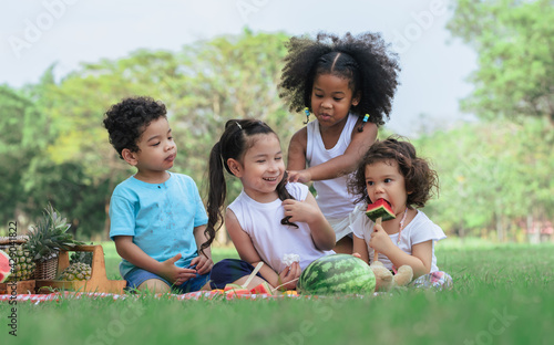 Group of four mixed race African and caucasian little cute kids sitting, playing in outdoor green park for picnic, eating fruit, watermelon, pineapple with freshness. Education and Diversity Concept. © Ann Rodchua
