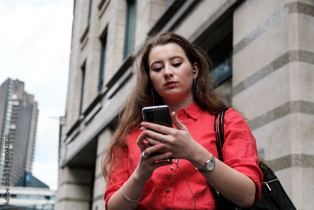 Young woman in a red shirt using her smartphone in the street, listen to the music with earphones.