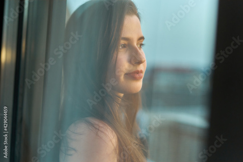 young woman with long hair and fair skin smiling through a window with the sunset in the background. teenager looking through a window. perfect smile pretty girl with unfocused city