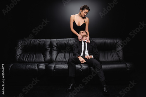 seductive woman in slip dress hugging man sitting on leather couch on black. © LIGHTFIELD STUDIOS