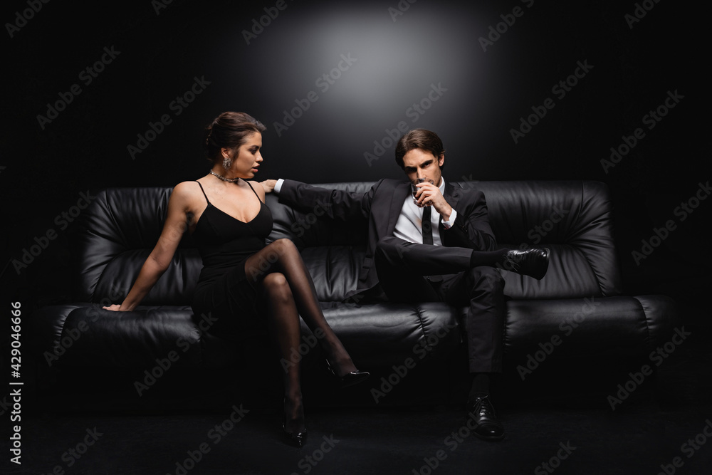 full length of man in suit sitting on leather sofa with glass of whiskey near woman in slip dress on black. full length of man in suit sitting on leather sofa with glass of whiskey near woman in slip dress on black.
