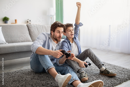 Father-son competition. Joyful boy winning dad in videogame and raising hand, celebrating victory photo