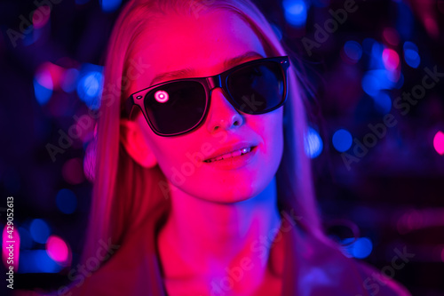 Young blond woman in sunglasses in a nightclub. The girl smiles and listens to moving music. Blurred background, close-up, red and blue lights. The concept of nightlife, parties and fun.