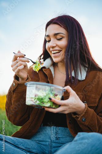 Laughing teenager girl enjoying eating a salad. She is in the field during sunset.