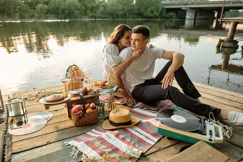 A young couple in love had a romantic picnic by the river at sunset. The girl gently hugs her young man.