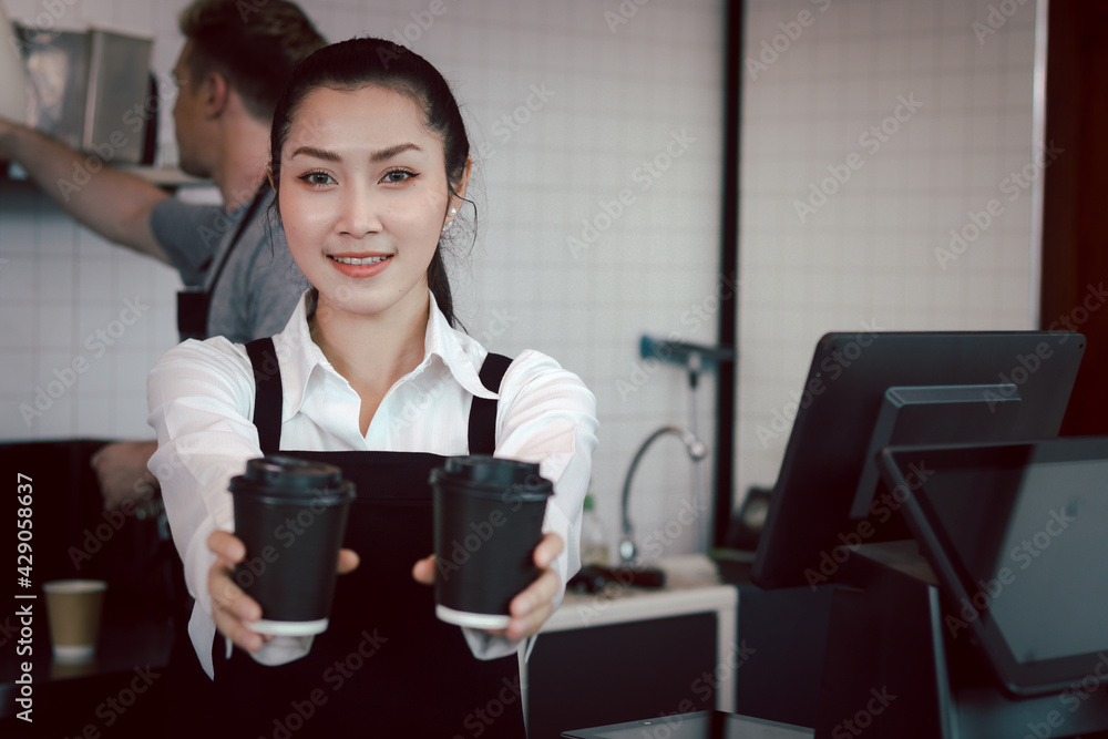 Young beautiful Asian female barista wearing apron, giving paper disposable coffee cups to camera while standing behind coffee shop counter bar, smiling waitress staff working at cafe. Young beautiful Asian female barista wearing apron, giving paper disposable coffee cups to camera while standing behind coffee shop counter bar, smiling waitress staff working at cafe.