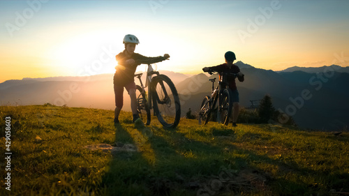 Mother and daughter walking uphill with mountain bike at a sunset.