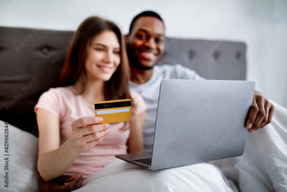 Cheerful young multiracial couple shopping on web from home, sitting on bed together, using laptop and credit card Cheerful young multiracial couple shopping on web from home, sitting on bed together, using laptop and credit card