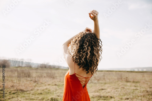 Young woman with curly hair, wearing a red summer backless dress, enjoying good weather, in a field. photo