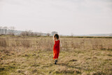 Happy young woman with curly hair, wearing a red summer dress outdoors, walking in a field.