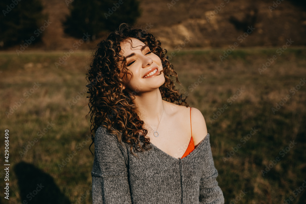 Portrait of a young woman with curly hair, smiling, outdoors, in sun light.