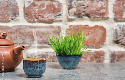 Hot green tea in a blue bowl, selective focus, steam rises above the cup, next to a clay teapot. Gray stone table, brick vintage background. Close-up, tea ceremony, minimalism, place for text