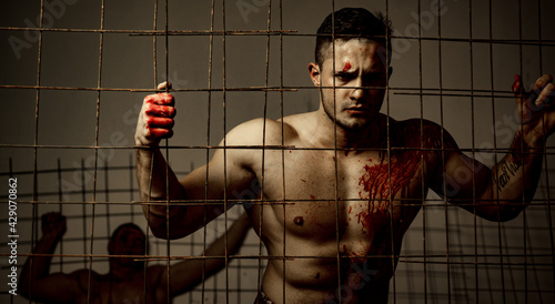 Strong aggressive monster behind grid. Prison for monster. Psycho mad man. Murderer mythical creature. Muscular man soiled with red blood. Halloween concept. Scary monster just murdered his victim