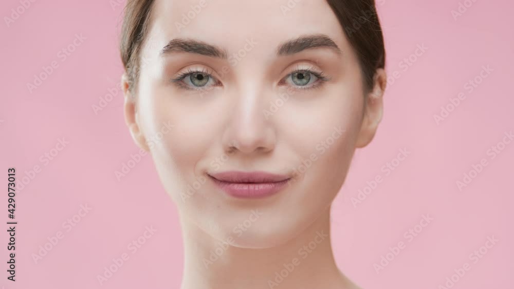 Good-looking fit Caucasian brunette girl with ponytail turns her head touching her face with hand, looking at the camera and smiling wide against pink ripple background | Skincare lotion commercial Good-looking fit Caucasian brunette girl with ponytail turns her head touching her face with hand, looking at the camera and smiling wide against pink ripple background | Skincare lotion commercial