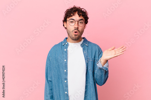 hispanic handsome man looking surprised and shocked, with jaw dropped holding an object with an open hand on the side photo