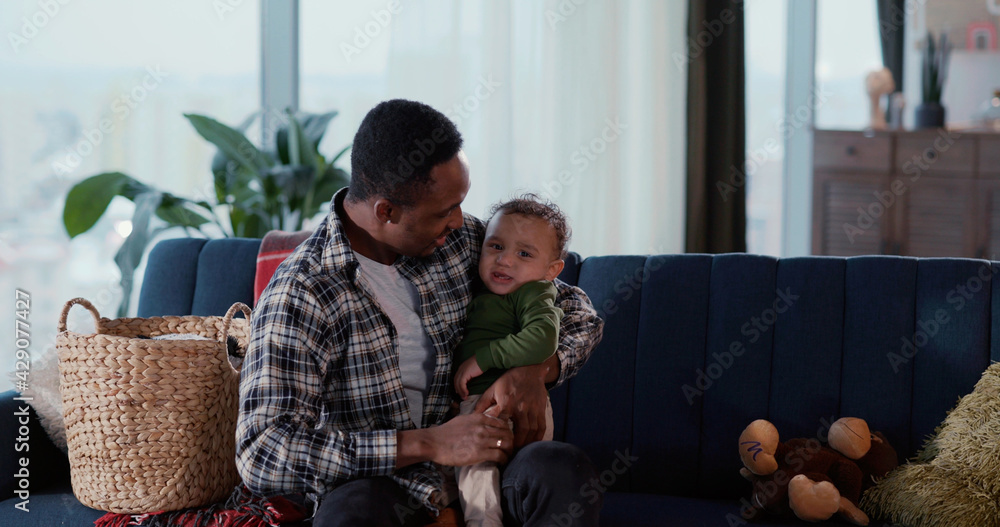 African american handsome happy man fatrher holding baby crying boy child on hands calming down together playing in living room. Family portrait.