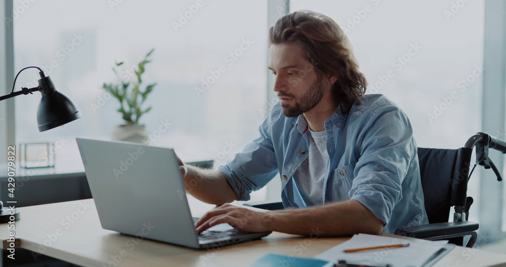 Young caucasian man disabled in wheelchar coming to office corporate workspace sitting by desk using computer laptop. Young caucasian man disabled in wheelchar coming to office corporate workspace sitting by desk using computer laptop.