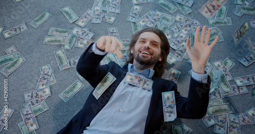 Young happy caucasian office worker lying on the floor cathcing dollar banknotes falling money rain. Wealthy lifestyle. Cute scene. Business success.
