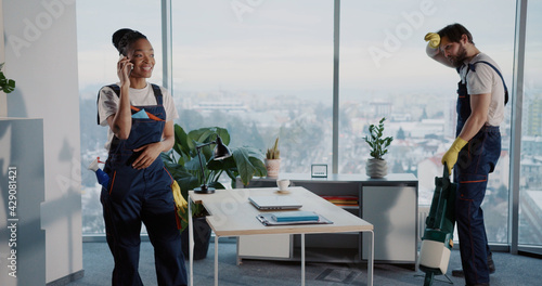Lazy african young talkative woman dialing mobile phone while her tired male colleague janitor cleaning office alone using vacuum cleanner.