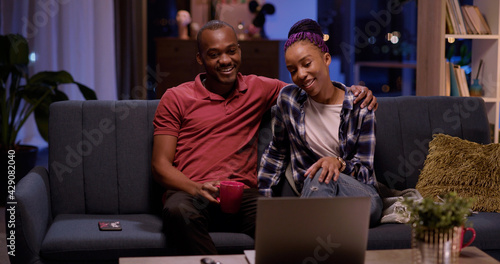 Young afro-american couple of attractive dating people staying home in cozy apartment, sharing news an drinks using laptop for work.