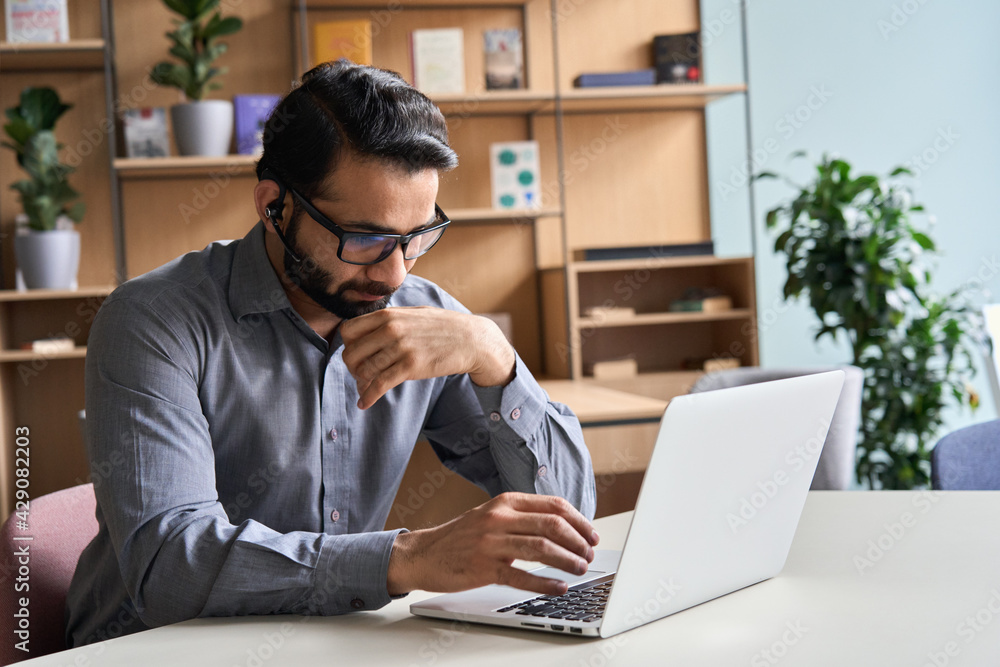 Latin indian serious businessman wearing glasses and headset having virtual team meeting call, remotely working at home watching online learning training webinar in remote office. Latin indian serious businessman wearing glasses and headset having virtual team meeting call, remotely working at home watching online learning training webinar in remote office.