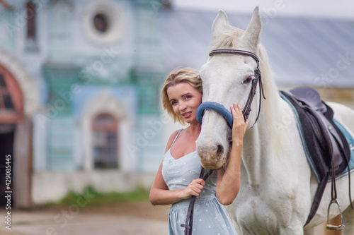 Beautiful woman in a blue dress hugs a white horse. Best friends. Photoshoot with a horse. Hobby love for animals. Country life. Romantic elegant girl tenderness. Saddle and bridle. Close-up portrait