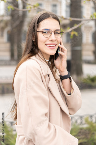 Charming young woman talking on the mobile phone wearing glasses and smiling outdoors in the city  © SlavaSelfStudio