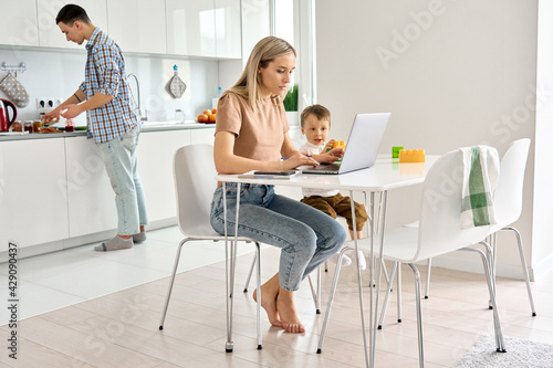 Busy young mother working from home office using laptop computer sitting at kitchen table with cute kid child son at home while dad cooking. Parents and child, distance virtual work in family life.