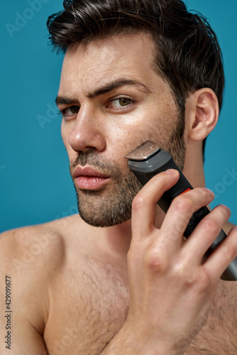 Closeup of brunette handsome guy looking at camera while using electric razor as a part of his morning routine isolated over blue background