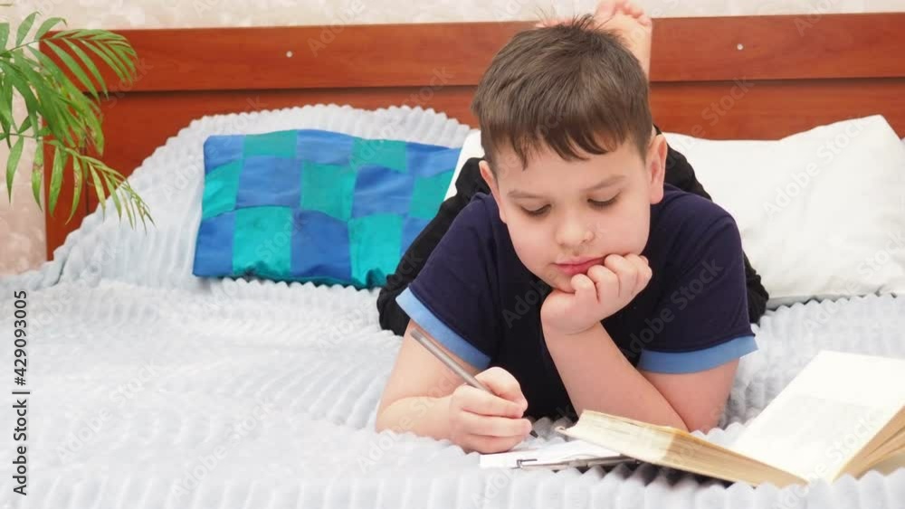 A boy reads a book while lying on the bed, does homework, writes, draws. Pensive little boy at home. Vacation and rest concept. Love for reading. Home schooling. Quarantine Holidays. Training courses A boy reads a book while lying on the bed, does homework, writes, draws. Pensive little boy at home. Vacation and rest concept. Love for reading. Home schooling. Quarantine Holidays. Training courses
