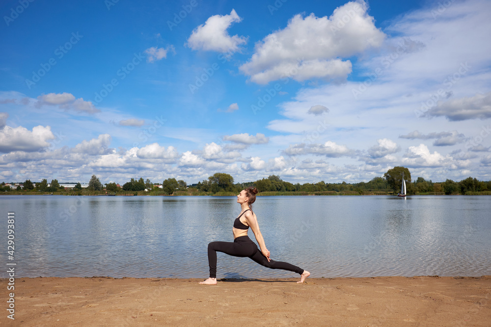 Fototapeta premium Young woman wearing black sportswear practicing yoga on the sand near lake.
