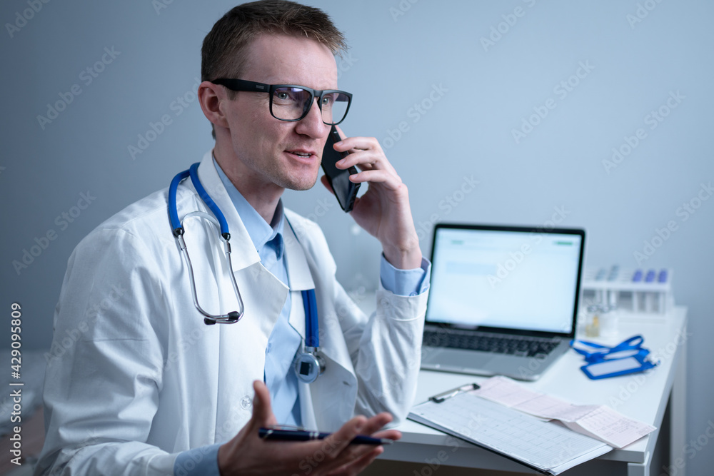 European doctor in white medical coat and glasses consults patient on cell phone in clinic office, sitting at table with laptop. German general practitioner at workplace in hospital talking on phone European doctor in white medical coat and glasses consults patient on cell phone in clinic office, sitting at table with laptop. German general practitioner at workplace in hospital talking on phone