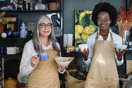 Two diverse saleswomen in beige apron and eyeglasses posing together at store an holding various cups in hands. Happy people working at decor shop.