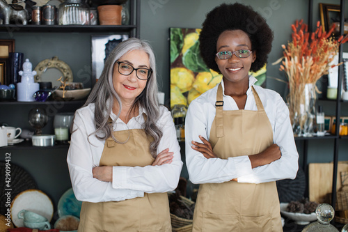 Front view of two multiracial saleswomen in beige aprons and eyeglasses smiling and posing with arms crossed at decor store. Concept of sale and service. photo