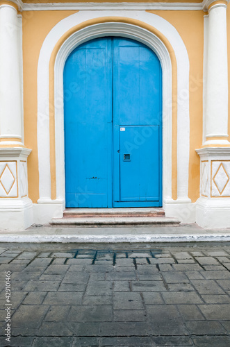 Close up of very large, tall arched bright blue wooden double doors with a smaller door cut into one set in a  yellow Spanish colonial house with decorative white columns, Campeche, Mexico © Compass Studios