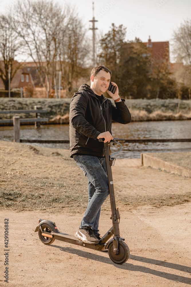 Obraz premium Man in black jacket drives an electric scooter outside. Explore the area with the electric scooter on vacation. Drive a scooter and talk on the phone. Use your mobile phone while driving 